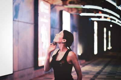 Young woman looking away while standing against wall at night