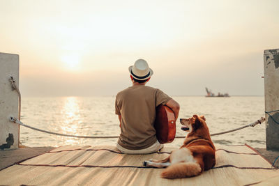 Rear view of man with dog sitting by sea against sky