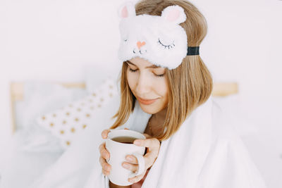 A young sick woman in pink pajamas and a sleeping mask on her head is sitting on the bed