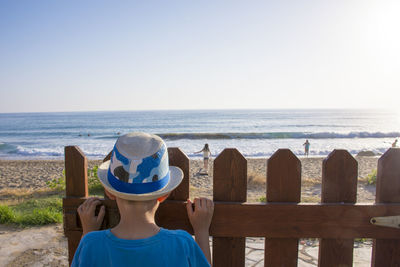 Rear view of boy standing by fence against beach