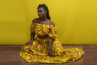 Young beautiful woman sitting on wooden floor, wearing colorful dress. against yellow background.