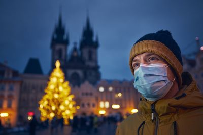 Close-up of man wearing mask standing against illuminated christmas tree at dusk