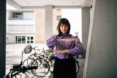 Portrait of young woman gesturing equal sign while standing by bicycle and wall