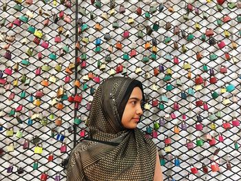 Thoughtful woman wearing hijab against colorful padlocks hanging on chainlink fence