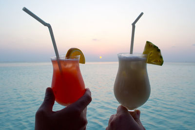 Close-up of hand holding drink against sea at sunset