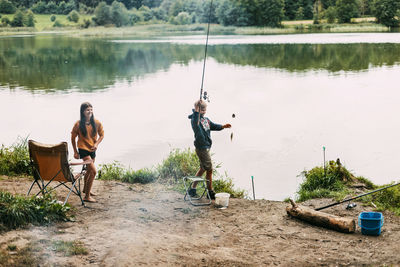 A brother and sister are standing on the river bank and fishing during a family vacation 