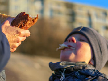 A boy is biting a piece from a loaf of bread which the heldin by his mother's hands.