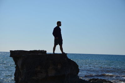 Silhouette man standing on rock by sea against clear sky