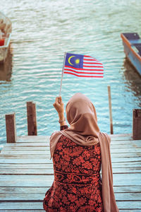 Rear view of woman wearing hijab holding malaysian flag sitting on pier by lake