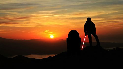 Silhouette hiker holding laser sword on mountain against sky during sunset