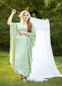 Pretty pregnant woman with big belly stands near clothesline with bed sheet, young girl takes care