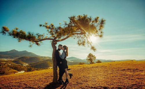 Side view of young couple kissing while standing on mountain against sky during sunny day