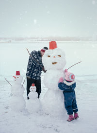 Father with daughter making snowman on field against sky
