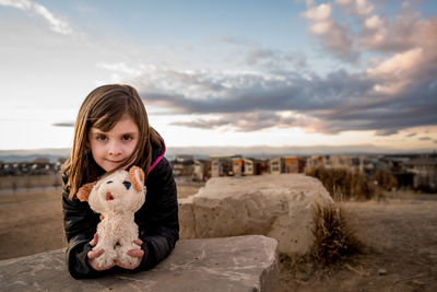 Young girl holds a stuffed dog at the top of a hill at sunset