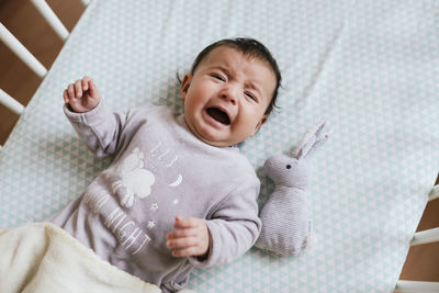 Portrait of crying baby girl lying in crib with toy bunny