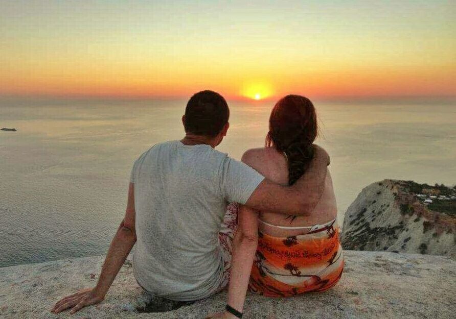 REAR VIEW OF FRIENDS SITTING ON BEACH AGAINST SUNSET SKY