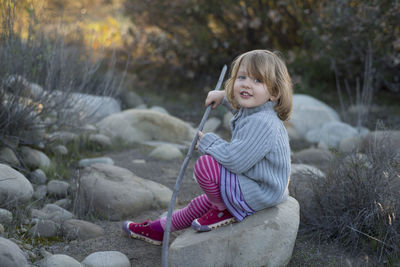 Portrait of girl holding stick while sitting on rock