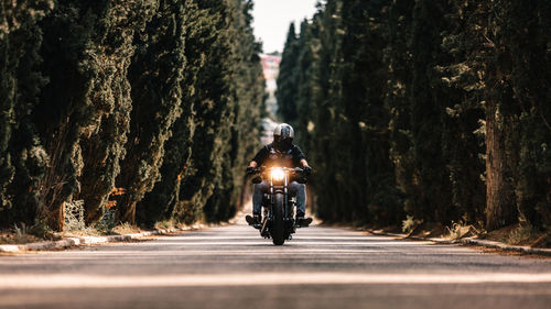 Biker in black leather jackets and helmet riding powerful motorcycle on asphalt road leading between green forest in countryside