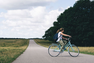 Side view of young woman cycling on road against sky