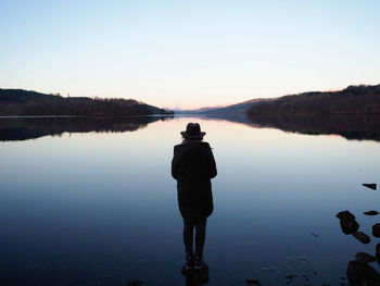 Rear view of silhouette man standing by lake against clear sky