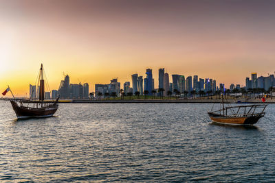 Sailboats in sea by buildings against sky during sunset