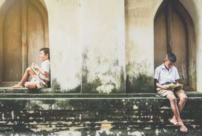 Full length of school boys sitting with books on retaining wall