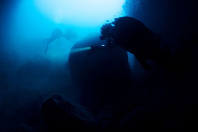 Professional divers with flashlight swimming near old sunken ship covered with moss on bottom of deep sea with clear water