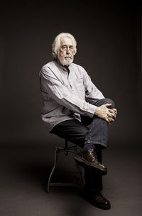 Portrait of senior man with white hair sitting on stool against black background