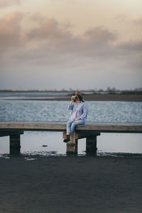 Woman photographing while sitting on pier against cloudy sky during sunset