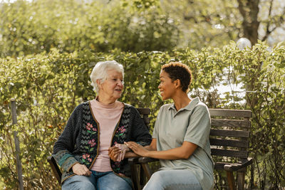 Smiling female caregiver talking with senior woman sitting on bench in park
