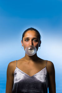 Portrait of serious young woman blowing bubble while standing against blue sky