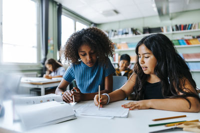 Female classmates writing while studying together and sitting near desk in classroom at elementary school