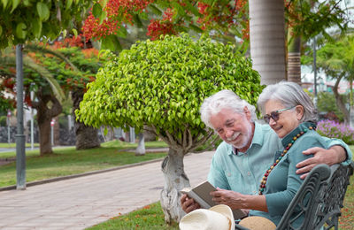 Smiling senior couple sitting on bench reading book at park