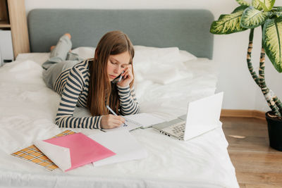 A happy girl is talking on the phone, lying in bed and using a laptop. online education, homework