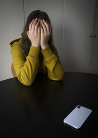 Sad teenager girl covered her face with hands while sitting at a table with a smartphone