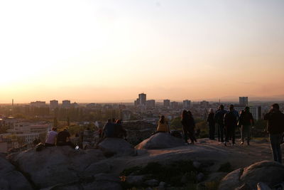 People on rock by buildings against sky during sunset