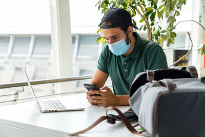 Smiling male wearing medical mask sitting at table with laptop and using cellphone