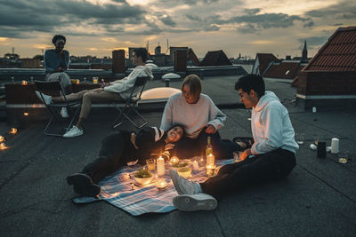 People sitting by bonfire in city against sky during sunset