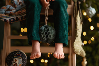 Barefoot boy in an elf costume holds craft paper deep green christmas tree ball