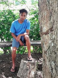 Full length portrait of young man sitting on tree trunk