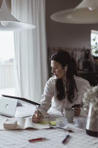 Businesswoman writing in diary while using digital tablet at home