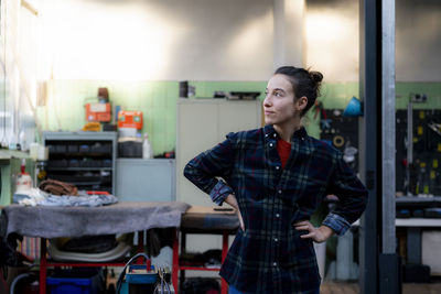 Female mechanic with hand on hip looking away while standing in workshop
