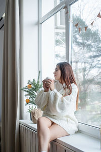 Woman in warm white winter sweater sitting on the window at home at christmas eve holding cu