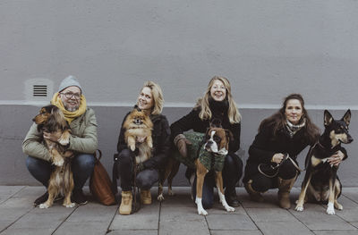 Portrait of smiling pet owners with dogs on footpath against wall in city