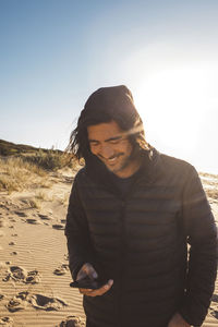 Smiling man using smart phone while standing at lakeshore against clear sky during sunset