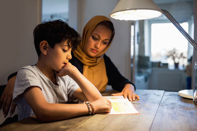 Mother and son reading book under illuminated desk lamp at home