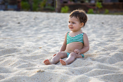 Adorable child in a swimsuit sits on a sandy beach in the sunshine.