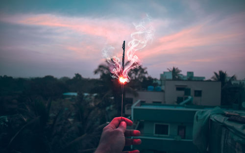 Cropped hand holding lit sparkler during sunset