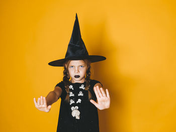 Portrait of girl wearing witch hat gesturing against yellow background during halloween