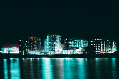 Illuminated buildings by sea against sky at night